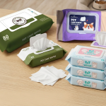 A variety of pet care wipes: a large 80-pack of olive green wipes, a purple pack with a cute corgi, and a stack of three smaller blue and pink wipes for puppies and kittens, arranged on a wooden table.